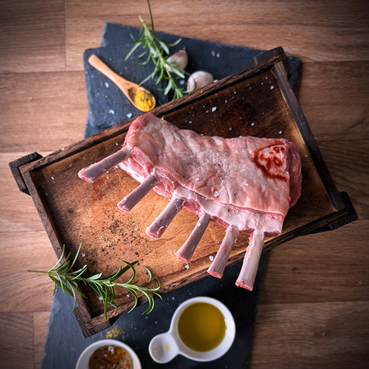 Raw lamb rack on a wooden tray with rosemary and a small bowl of oil on a wooden surface.