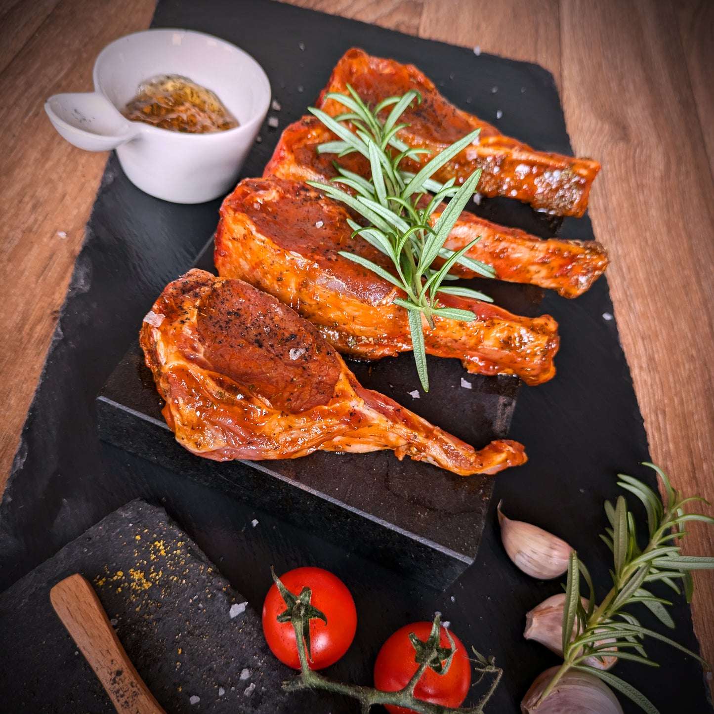 Roasted lamb chops on a black slate board with herbs and tomatoes on a wooden surface